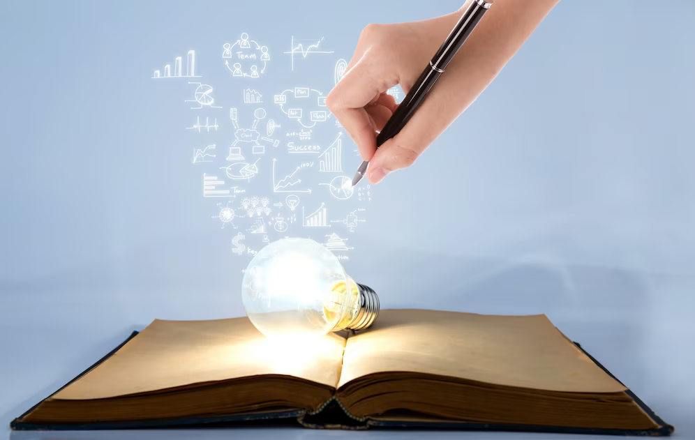 Im unteren Teil des Bildes ist ein aufgeklapptes Buch zu sehen, auf welchem eine leuchtende Glühbirne liegt, aus der Symbole herausstrahlen. Oben ist eine Hand zu sehen, die mit einem Stift an den Symbolen malt