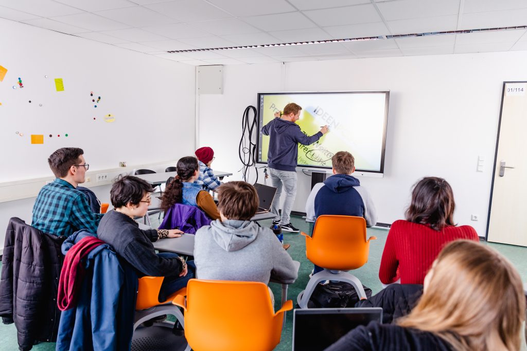 Ein Seminarraum, in dem ein Dozent vorne auf einem digitalen Whiteboard schreibt. Hinter ihm sitzen acht Studierende, die zu ihm nach vorne schauen.