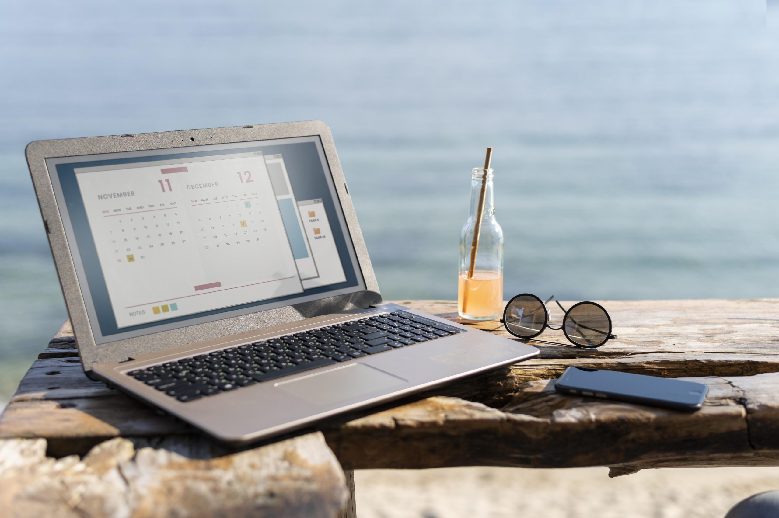 Laptop, Trinkflasche, Sonnenbrille und Handy liegen auf einem Holztisch, im Hintergrund ist das Meer zu sehen.